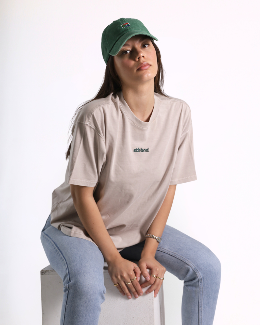 A woman wearing a beige short-sleeve t-shirt with a small black 'athlet' logo on the chest, paired with light blue jeans and a green baseball cap, sitting on a white block against a white background.