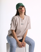 A woman wearing a beige short-sleeve t-shirt with a small black 'athlet' logo on the chest, paired with light blue jeans and a green baseball cap, sitting on a white block against a white background.