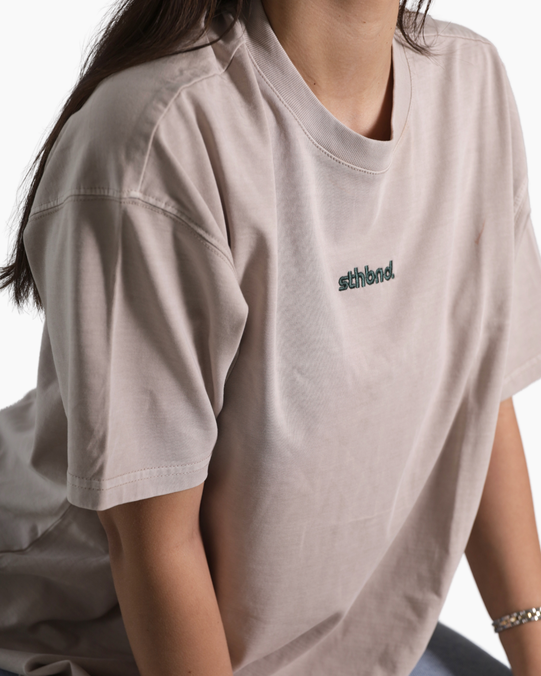 A close-up of a person wearing an oversized light taupe t-shirt with a small embroidered 'sfbird.' logo on the left chest.