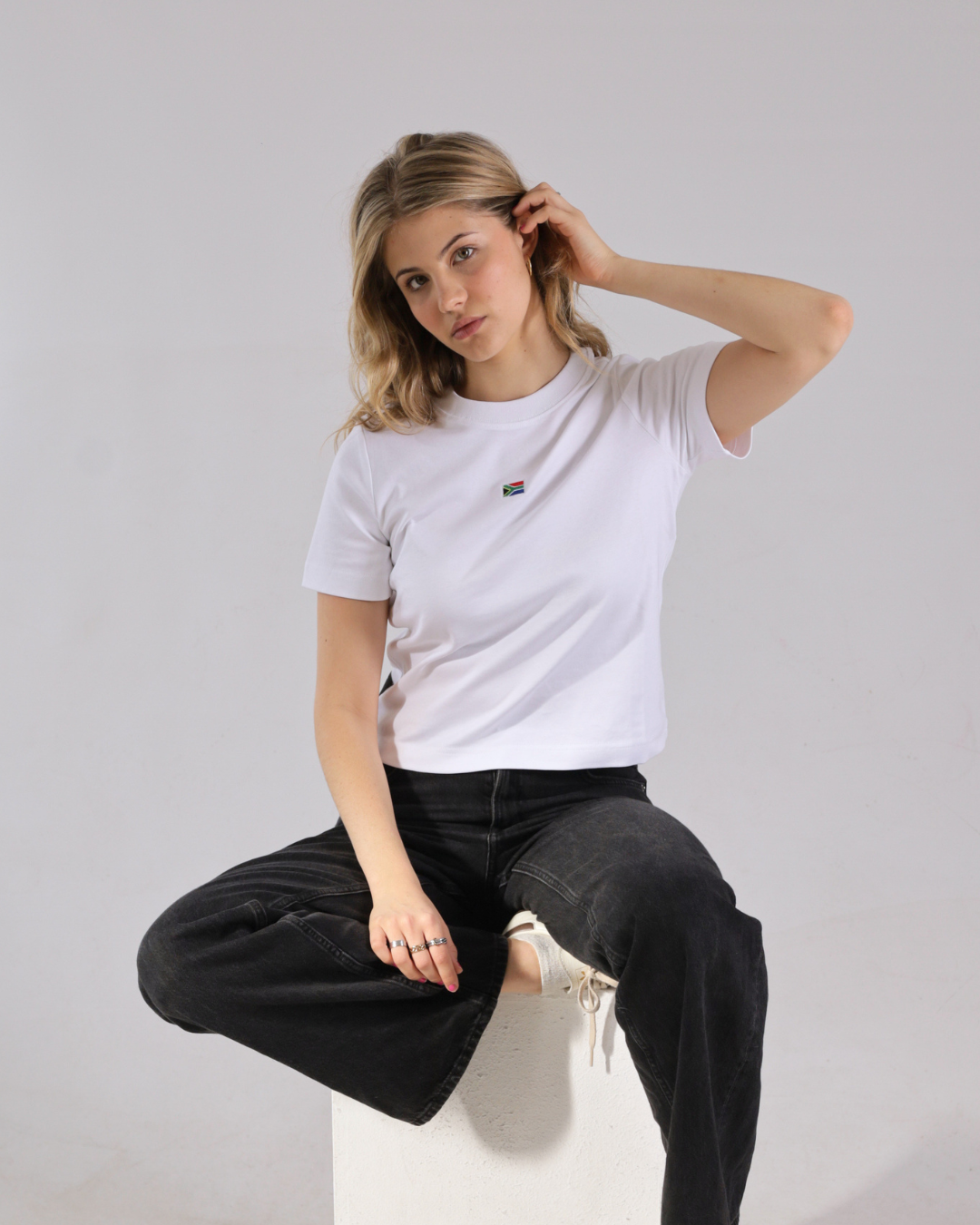 A woman sitting on a white block wearing a white short-sleeve t-shirt with a small rainbow flag graphic on the left chest, paired with black jeans.
