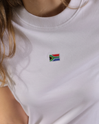 A close-up of a person wearing a white crewneck t-shirt with a small embroidered South African flag patch on the left chest.