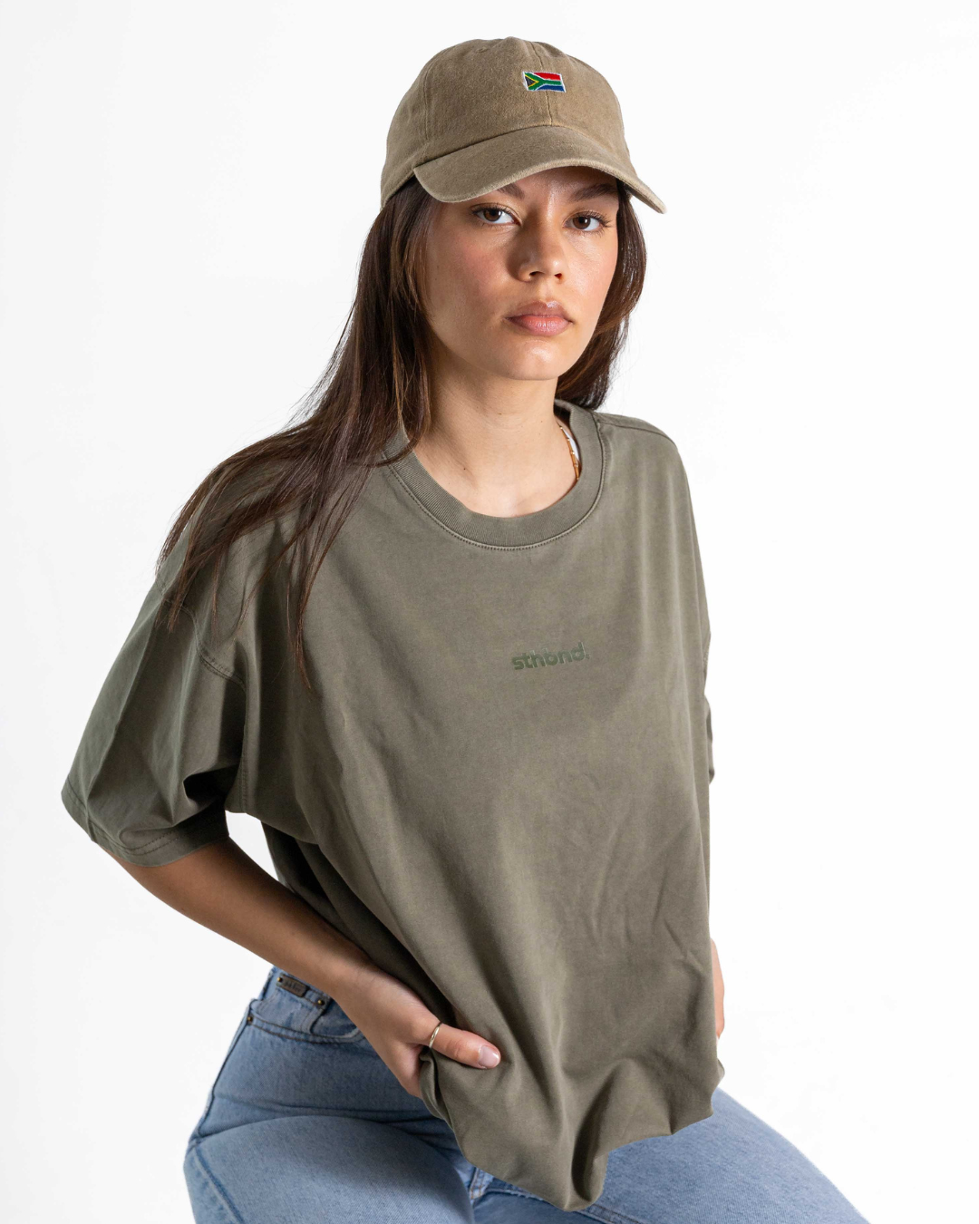 A woman wearing an oversized olive green t-shirt with a small green 'abound.' logo on the chest, paired with a matching olive green baseball cap and blue jeans, photographed against a clean white background.
