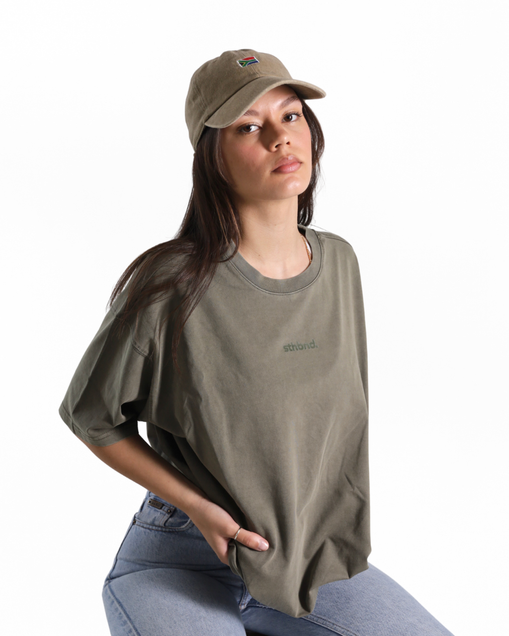 A woman wearing an oversized olive green t-shirt with a small green logo on the chest, paired with a matching olive green baseball cap and light blue jeans, photographed against a solid white background.