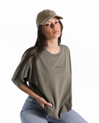 A woman wearing an oversized olive green t-shirt with a small green logo on the chest, paired with a matching olive green baseball cap and light blue jeans, photographed against a solid white background.