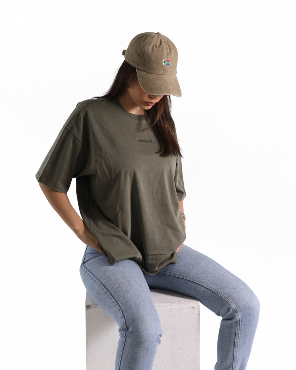 A woman wearing an oversized olive green t-shirt, light blue jeans, and a khaki baseball cap sits on a white block against a white background.