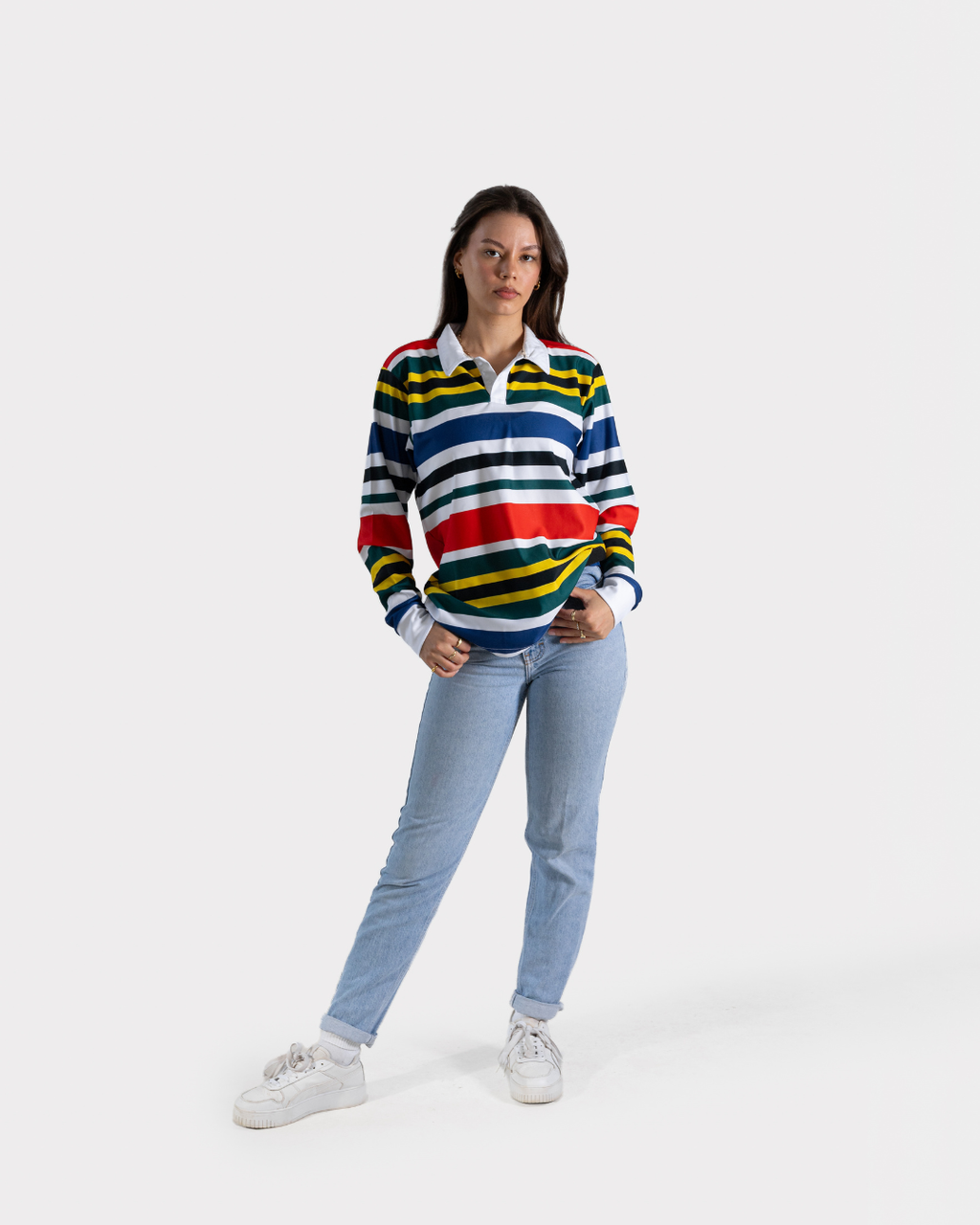 A woman modeling a long-sleeve rugby-style striped sweater with blue, white, yellow, and red horizontal stripes, paired with light blue jeans and white sneakers, against a light gray studio background.