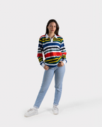 A woman modeling a long-sleeve rugby-style striped sweater with blue, white, yellow, and red horizontal stripes, paired with light blue jeans and white sneakers, against a light gray studio background.