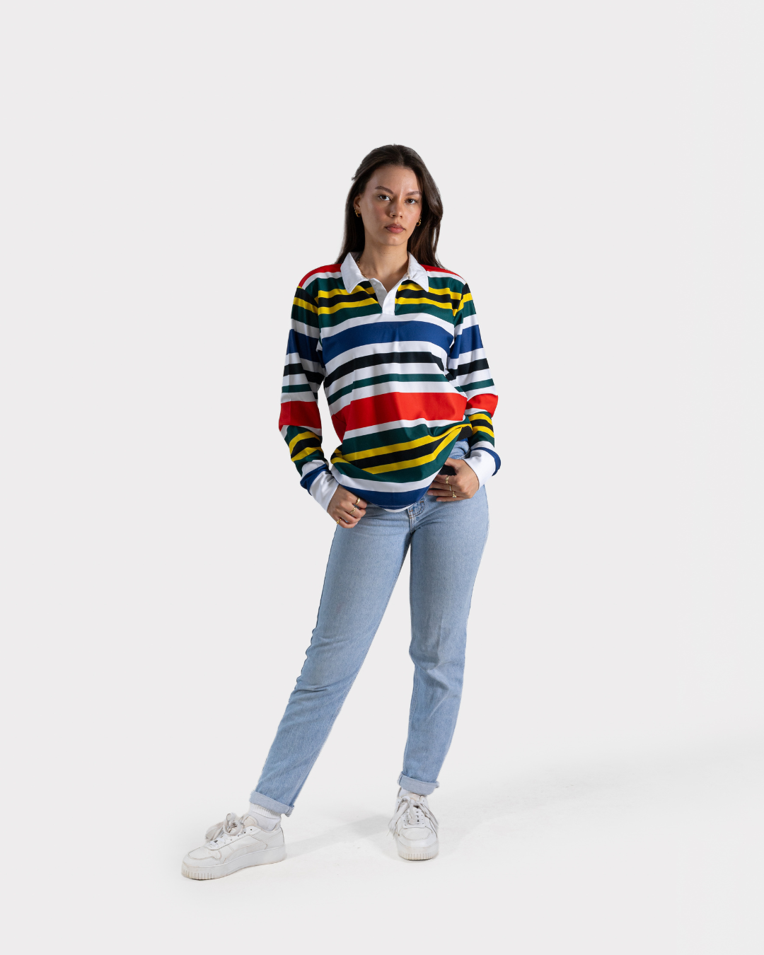 A woman modeling a long-sleeve rugby-style striped sweater with blue, white, yellow, and red horizontal stripes, paired with light blue jeans and white sneakers, against a light gray studio background.