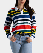 A woman wearing a long-sleeve rugby-style striped shirt with horizontal bands of red, green, blue, white, black, and yellow, paired with light blue jeans against a light gray background.