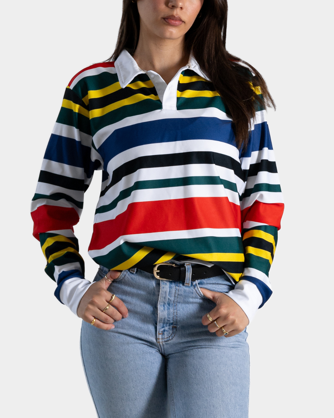A woman wearing a long-sleeve rugby-style striped shirt with horizontal bands of red, green, blue, white, black, and yellow, paired with light blue jeans against a light gray background.