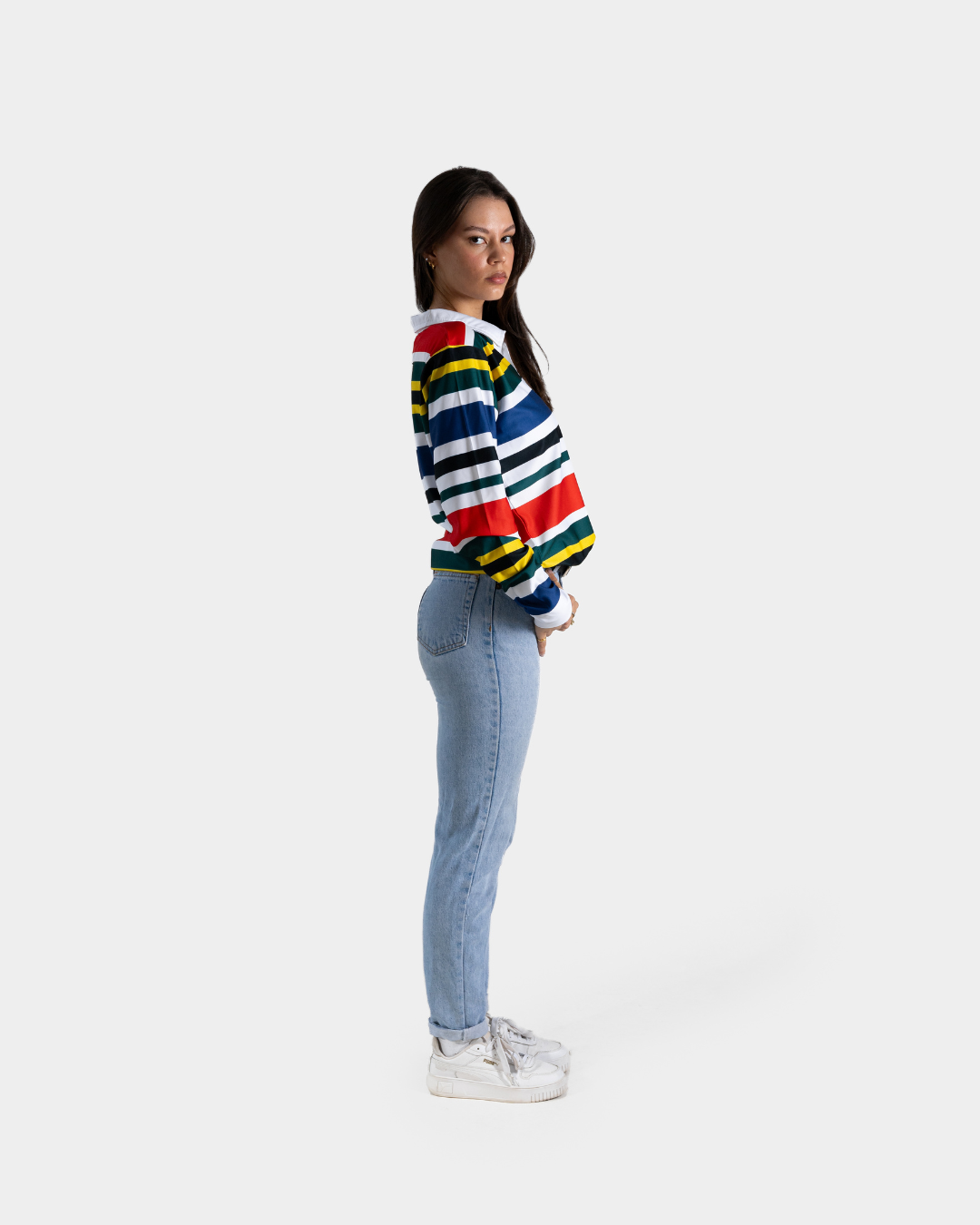 A woman standing in profile wearing a colorful striped knit sweater with blue jeans and white sneakers, photographed against a plain light gray studio background.