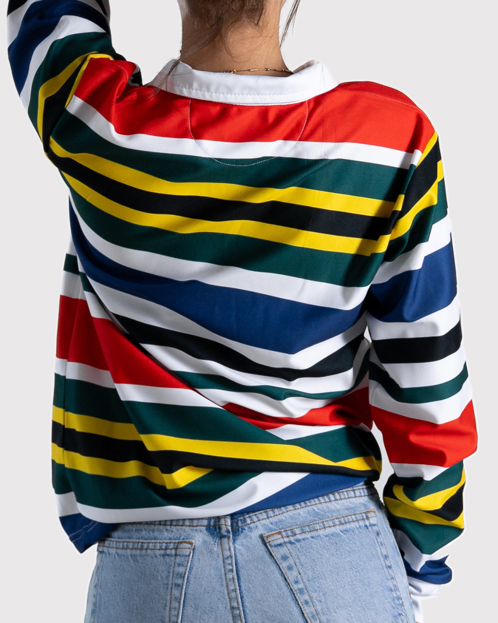 A person shown from the back wearing a long-sleeve collared top with bold horizontal multicolor stripes in red, green, blue, yellow, black, and white, paired with light blue jeans against a light gray studio background.