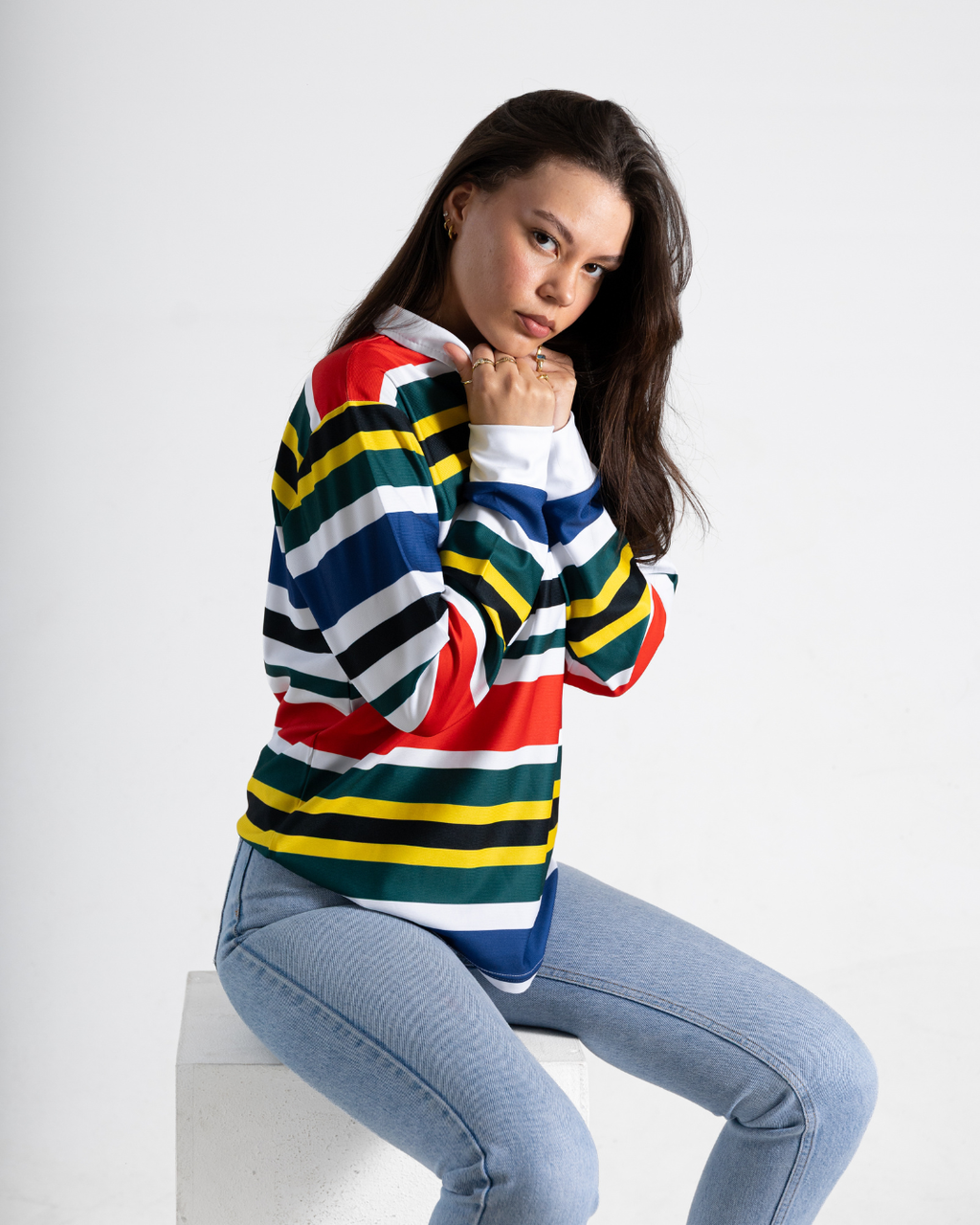A woman wearing a long-sleeve striped sweater with horizontal bands of red, yellow, green, white, and blue, paired with light blue jeans, sitting on a white block against a light gray background.