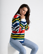 A woman wearing a long-sleeve striped sweater with horizontal bands of red, yellow, green, white, and blue, paired with light blue jeans, sitting on a white block against a light gray background.
